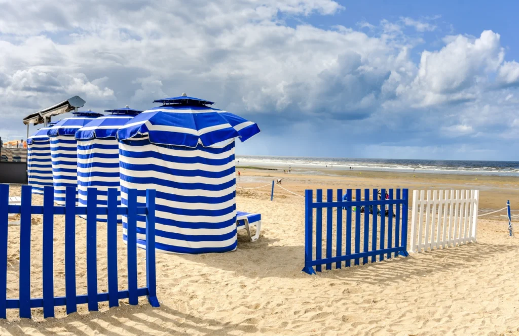 Sur la plage de Cabourg, tentes de plage rayées bleues et blanches sur un sable fin, avec la mer à l'horizon et un ciel nuageux.